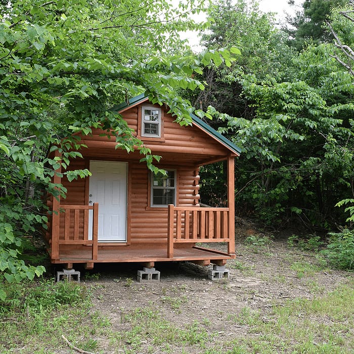 A small wooden cabin with a front porch stands elevated on concrete blocks, surrounded by dense green foliage. The cabin has a white door and a small window above it with a gable roof. The setting appears to be a peaceful, forested area.
