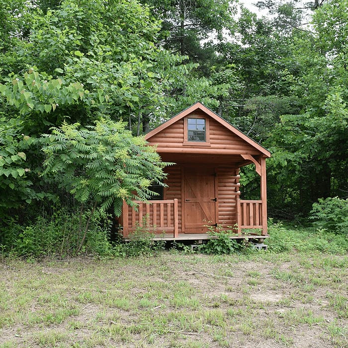 A small wooden cabin with a porch stands surrounded by dense green foliage and trees. The cabin, which has a front door and a window in the upper loft, is slightly raised off the ground. The area in front of the cabin is a cleared grassy patch.