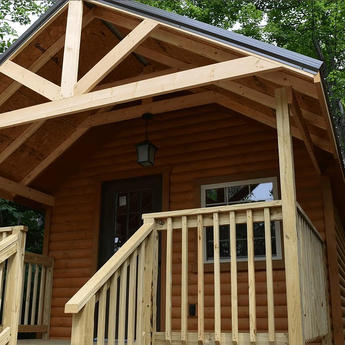A small, rustic log cabin with a wooden porch and stairs nestled in a lush forest setting. The cabin features exposed wooden beams, a gabled roof, and a hanging lantern next to the front door. Dense green foliage surrounds the cabin.