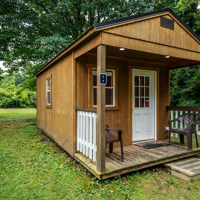 A small wooden cabin with a covered porch stands in a grassy area surrounded by trees, offering charming lodging options. The porch has two plastic chairs and a small white railing. A picnic table can be seen in the background to the left. The cabin's door has a window and a welcome mat in front.