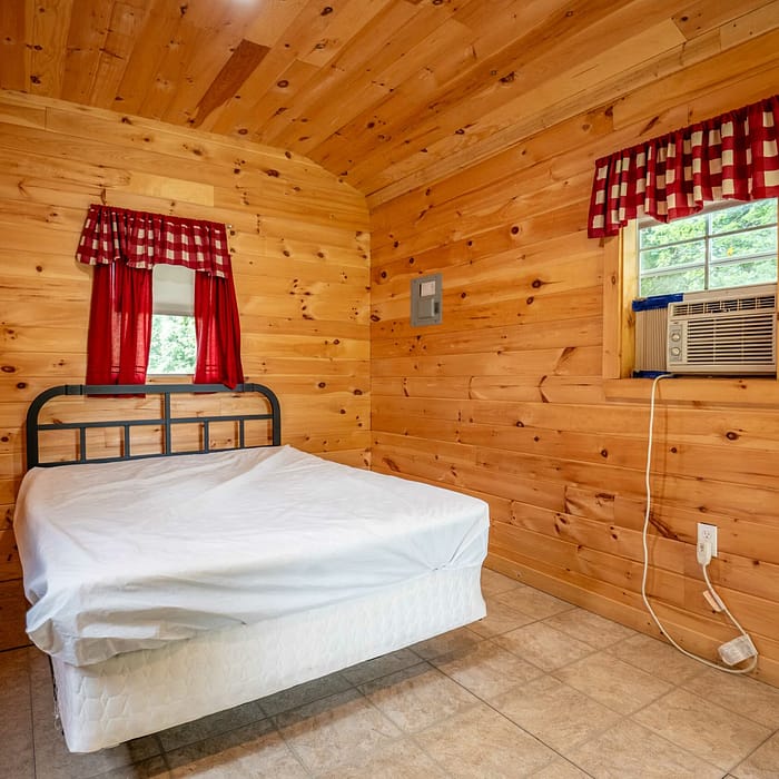 A simple cabin bedroom with wooden walls and a tiled floor features a metal-framed bed with a white mattress but no bedding. Two windows with red and white checkered curtains brighten the accommodations. An air conditioning unit is installed in one window.
