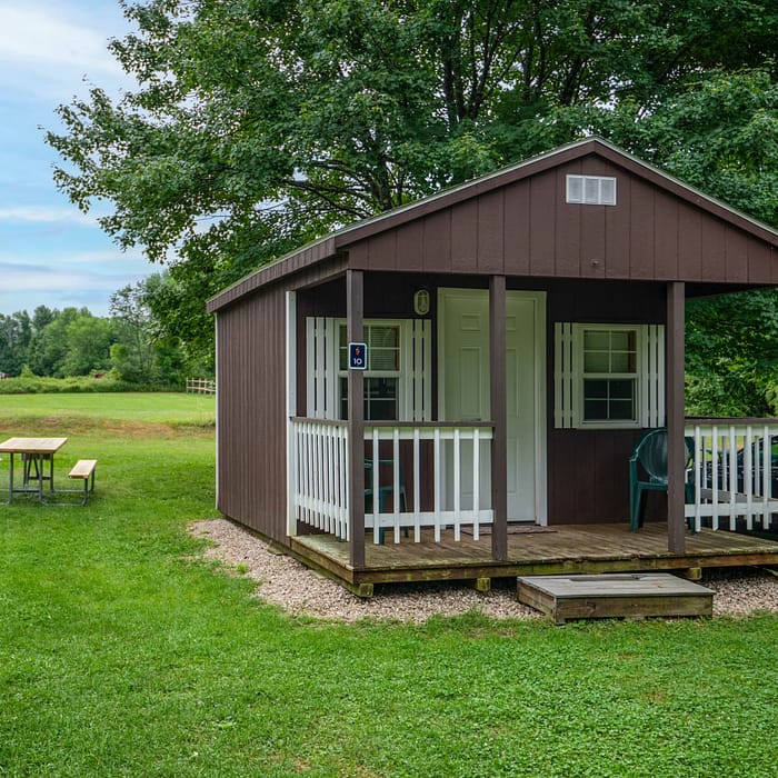 A small, brown wooden cabin with white trim and a front porch sits on a grassy lawn. This cozy lodging features a black car parked nearby and a wooden picnic table on the left side of the cabin. Surrounded by trees and a large open field under a blue sky with light clouds, it’s an idyllic place to stay.