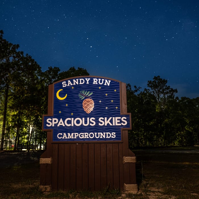 A wooden sign reads "Sandy Run, Spacious Skies Campgrounds" with a pineapple and crescent moon design. It stands in a wooded area under a starry night sky. Trees are visible in the background.