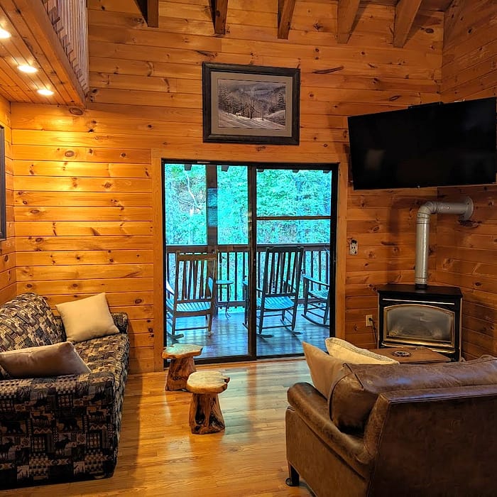A cozy wooden cabin interior features a seating area with two couches and a small table. A fireplace and TV are mounted on the wall. A sliding glass door opens to a balcony with rocking chairs, overlooking a lush, wooded area.