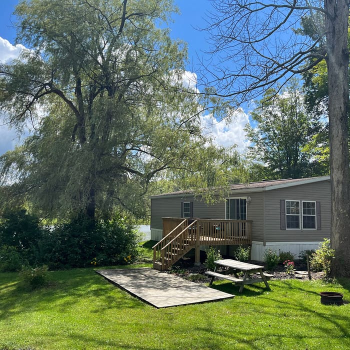 A small gray house with a wooden deck sits by a lake surrounded by trees. The yard has a picnic table, a fire pit, and a small path. The sky is blue with scattered clouds, and sunlight illuminates the green grass.