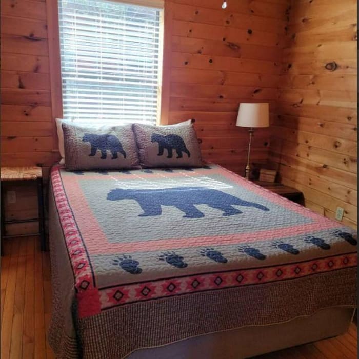 Cozy rustic bedroom with wooden walls and ceiling. Bed adorned with a quilt featuring bear designs and matching pillows. A window with blinds lets in natural light. A small side table with a lamp is next to the bed. Hardwood floor adds warmth.