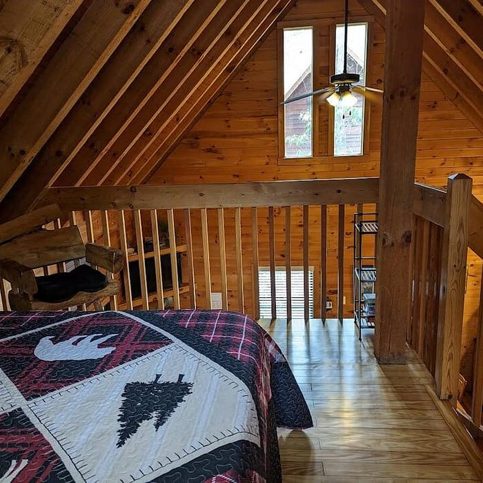 A cozy loft bedroom in a rustic cabin featuring wooden walls and ceiling. The room includes a bed with a patterned quilt, a small window above the headboard, and a ceiling fan. Natural light streams through a triangular window.