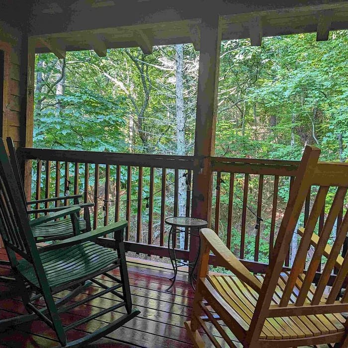 Two rocking chairs, one green and one light wood, are on a wooden porch surrounded by a forest of lush green trees. A small round table is placed between the chairs. The scene is serene and inviting, offering a peaceful view of nature.