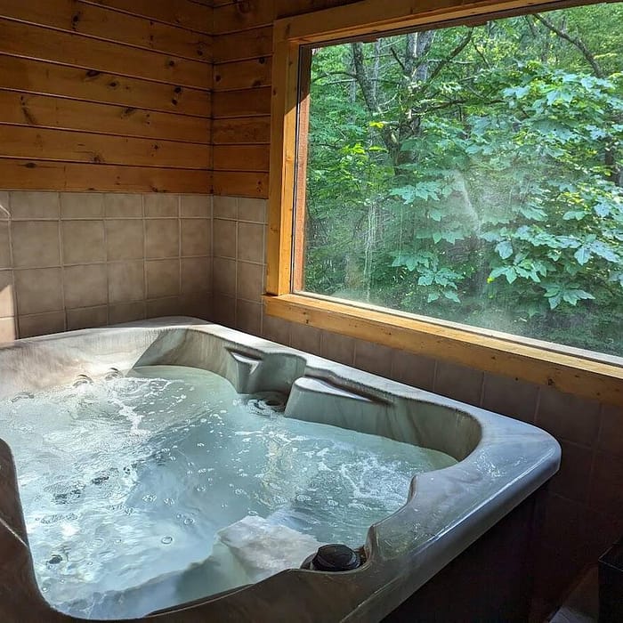 A cozy indoor hot tub with bubbling water is set in a wooden cabin room. A large window offers a view of lush green trees under a bright sky, creating a relaxing and natural atmosphere.