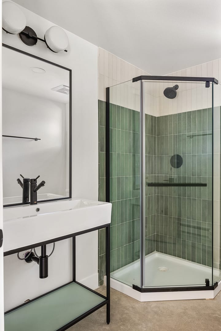 A modern bathroom with a glass-enclosed corner shower featuring green vertical tiles, a white rectangular sink with a black faucet, a black-framed mirror, and a minimalist vanity with an open shelf.