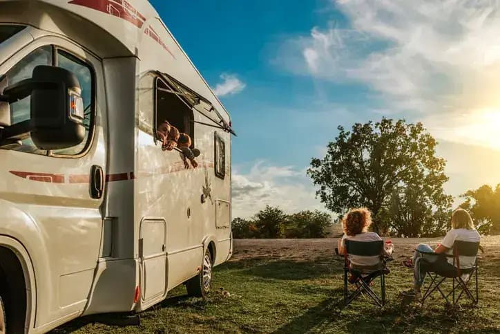 Two people sit in camping chairs outside a white RV parked on a grassy area, soaking in the beauty of one of the best New England RV sites. One person leans out of an RV window, smiling at them. A large tree and a vibrant, partly cloudy sky are in the background with the sun casting a warm glow.