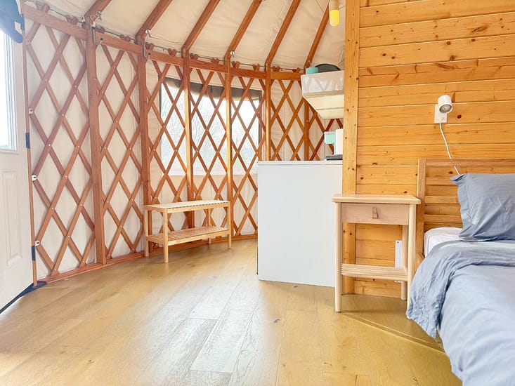 Interior of a bright yurt with wooden walls, flooring, and furniture, featuring a bed and natural light.