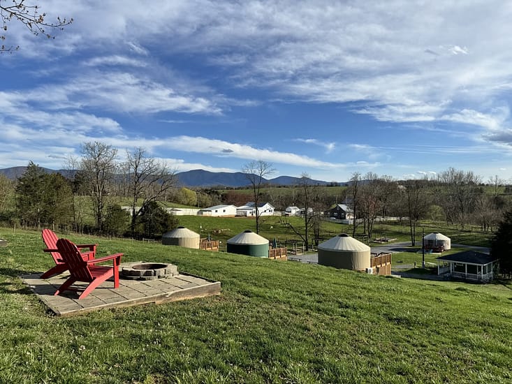 Rural landscape with yurts, a fire pit, and red chairs overlooking mountains.