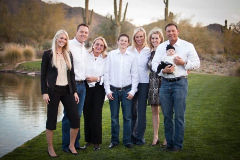 Eight people, including a baby, stand together outdoors on grass near water and desert plants, all dressed in semi-formal attire and smiling at the camera.