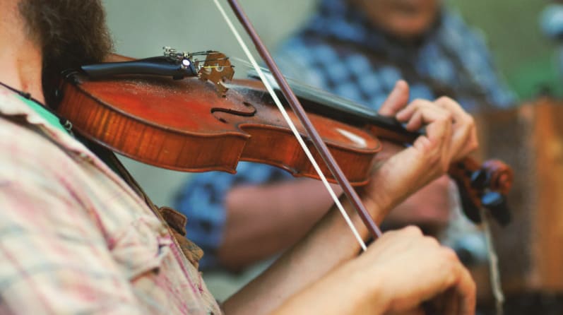 Close-up of a person playing a violin with a bow, focusing on the hands and instrument. Another person in a plaid shirt appears blurred in the background.