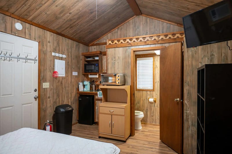 A small rustic room with wooden walls and ceiling. The space includes a kitchenette with a microwave, toaster oven, coffee maker, and mini-fridge. A door leads to a bathroom with a visible toilet. A mounted flat-screen TV is on the wall near the ceiling.