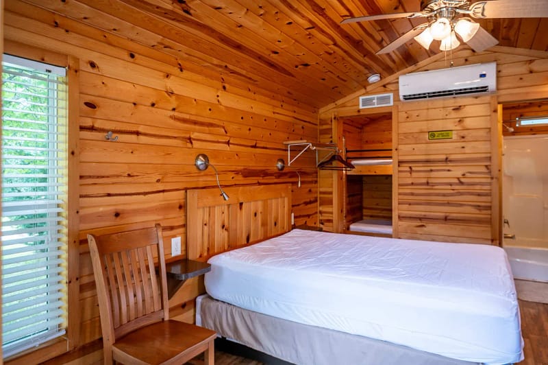 A cozy cabin room with wood-paneled walls and ceiling. It contains a double bed with white bedding, a wooden chair next to a window with blinds, a ceiling fan, AC unit, and an open closet with hangers. A bathroom is visible in the background.