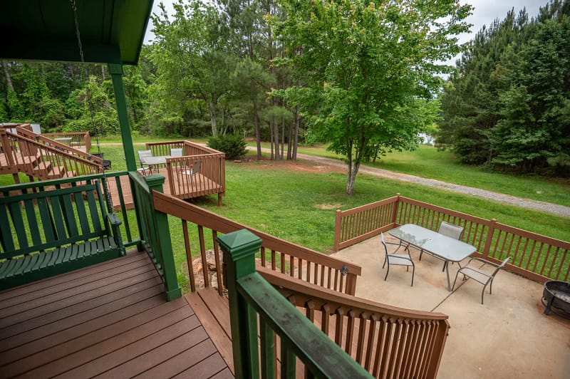 A scenic view of a backyard with multiple elevated wooden decks and stairways overlooking a lush green lawn and forested area. There is outdoor furniture, including a glass-top table with chairs on one of the patio spaces.