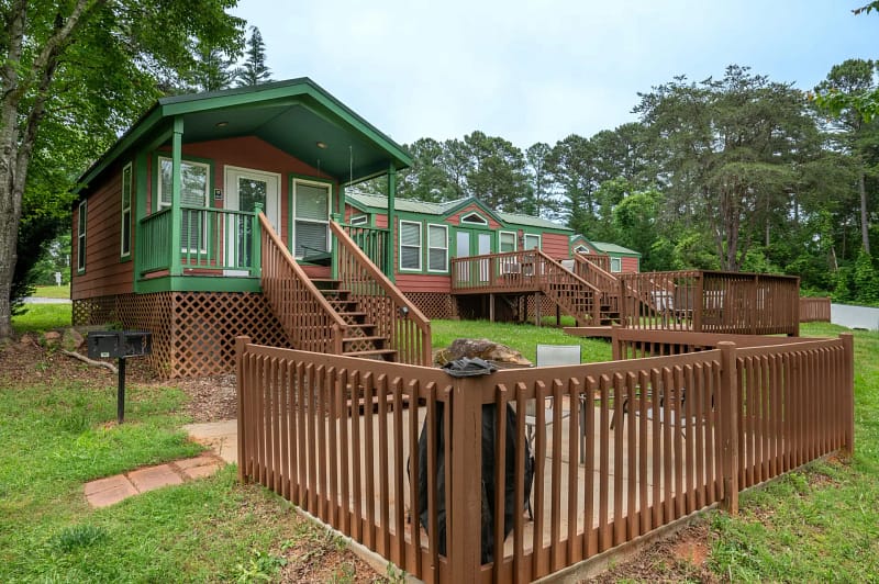 A row of small, elevated cabins with green roofs and brown exteriors, each with a wooden porch and steps leading to the front door. A fenced patio area with a grill and seating is in the foreground, surrounded by lush trees and greenery.