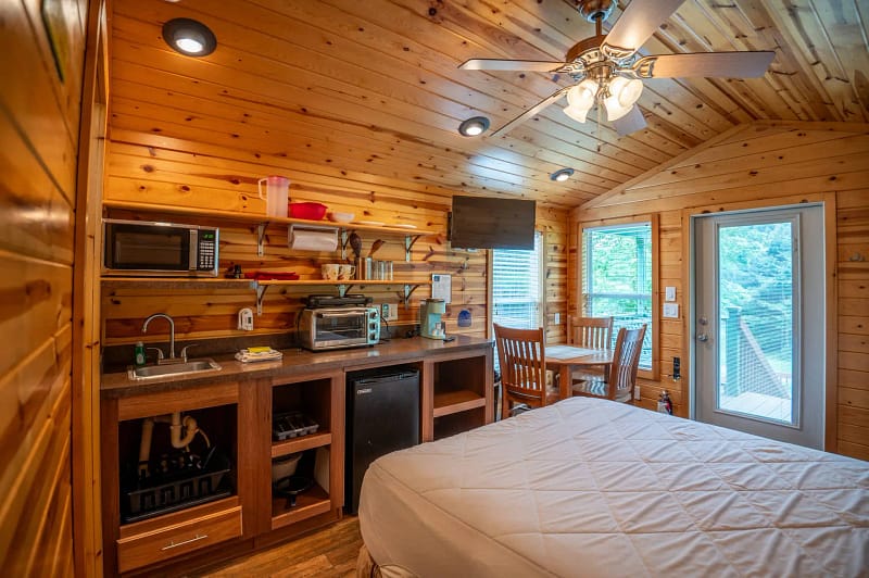 A cozy cabin interior with wooden walls and ceiling, featuring a kitchenette with a microwave, toaster, sink, and mini-fridge. A small dining table with two chairs is placed near a window and a door leading outside. A ceiling fan with lights is overhead.