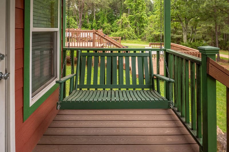 A green wooden porch swing attached to the railing of a wooden deck on a red house with green trim. The background features a lush green lawn, trees, and a raised wooden deck with stairs leading down to the grassy area.