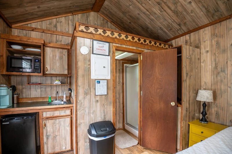 A cozy wooden cabin interior featuring a small kitchenette with a mini-fridge, microwave, coffee maker, and sink. A door leading to a bathroom is open, and a waste bin is in the foreground. A yellow nightstand with a lamp is beside a bed, not fully visible.