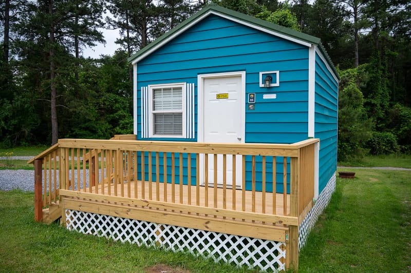 A small blue house with white trim and a front porch. The porch has a wooden railing and lattice skirting. The house has one window with shutters next to a white door. The surrounding area is grassy with trees in the background.