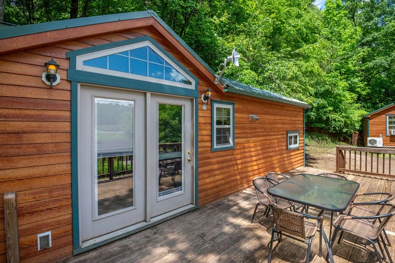 A wooden cabin with a green roof features a set of double glass doors, a small window, and a deck. The deck has an outdoor table and chairs. In the background, another similar cabin is visible, and the area is surrounded by lush green trees.