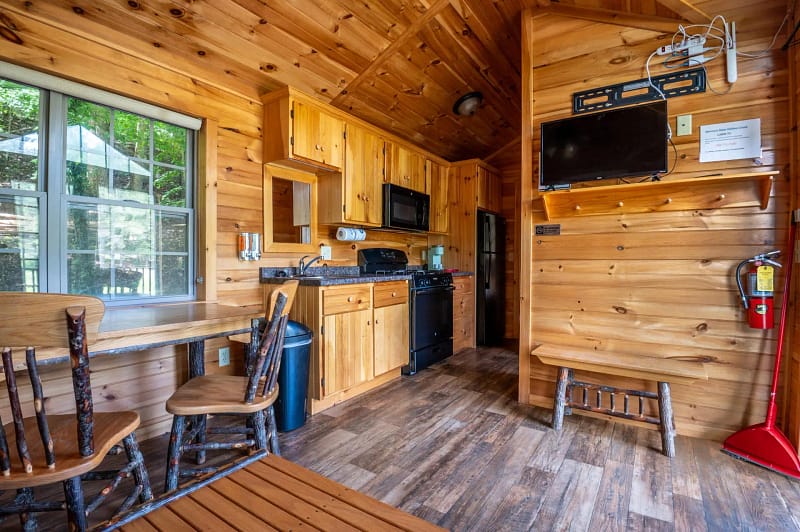 A cozy wooden cabin interior with a small kitchen equipped with a stove, oven, microwave, sink, and cabinets. A wall-mounted TV is above a small bench. Adjacent is a dining area with a rustic table and four chairs. A window to the left allows natural light to enter.