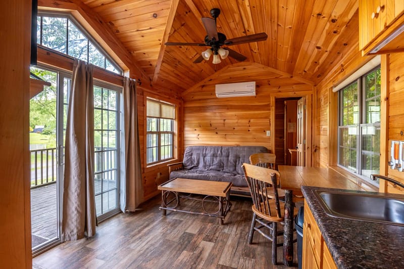 A cozy wooden cabin interior with large windows, a ceiling fan, and a futon sofa. The space is illuminated by natural light, featuring a small wooden coffee table, chairs, and a kitchenette with a counter and sink on the right side. A scenic view of greenery is visible outside.