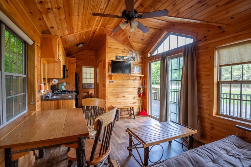 A cozy cabin interior with wooden walls and ceiling. The room features a dining table with four chairs, a kitchenette with an oven and microwave, a flat-screen TV mounted on the wall, a sofa, and large windows with a view of the outdoors. There is a ceiling fan above.