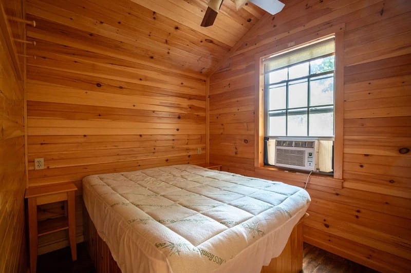 A small wooden cabin bedroom with a ceiling fan. The room features wood-paneled walls and ceiling, a window with an air conditioning unit, a double bed with white bedding, and a small wooden table in the corner. The window allows natural light into the room.