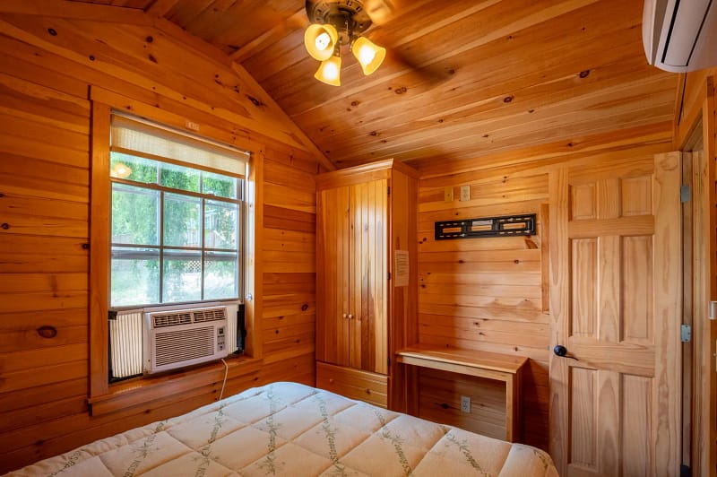 A cozy wooden cabin bedroom with a queen-sized bed featuring a patterned mattress. The room has wooden walls and ceiling, a ceiling fan, an air conditioning unit under the window, a wooden wardrobe beside a small wooden desk, and a wooden door. Warm lighting illuminates the space.