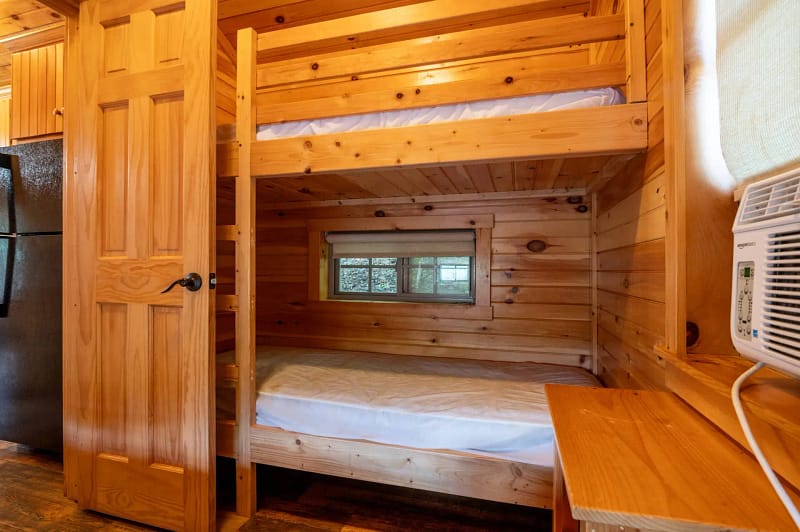 A cozy room features wooden bunk beds against a paneled wall. A small window is situated between the beds, letting in natural light. An air conditioner is mounted on the wall to the right, and a refrigerator is partially visible on the left.