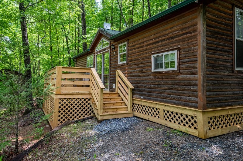 A small log cabin surrounded by lush green forest. The cabin has a wooden exterior, a green roof, and several windows. A wooden deck with railings and steps leads up to the front door. The area around the cabin is covered with gravel and greenery.