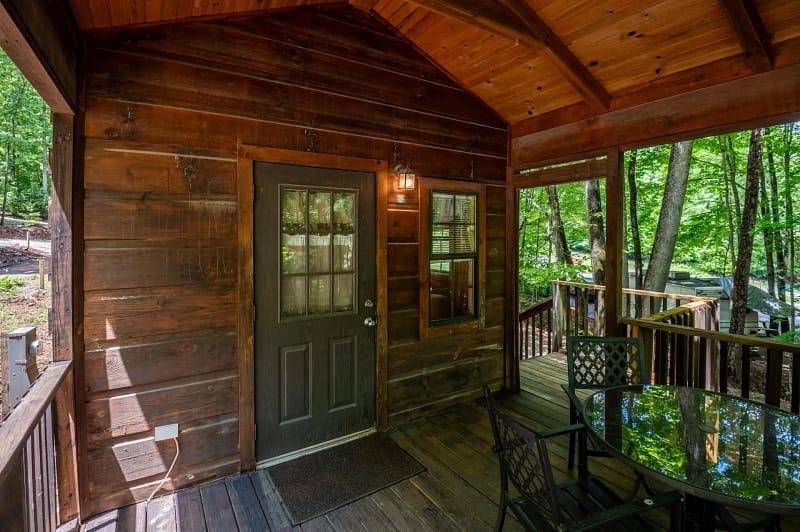 A wooden cabin porch featuring a door with window panes and a lit sconce above. Adjacent to the door is a window. The porch has a wooden ceiling and flooring, with a round glass table and four metal chairs in view. Surrounding trees are visible in the background.