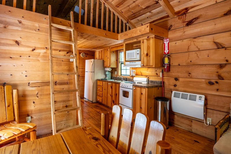 A cozy wooden cabin interior with a small kitchen. The kitchen includes a refrigerator, microwave, oven, and sink. There is a wooden dining table and chairs, a loft accessed by a ladder, a heater on the wall, and a fire extinguisher mounted near the ceiling.