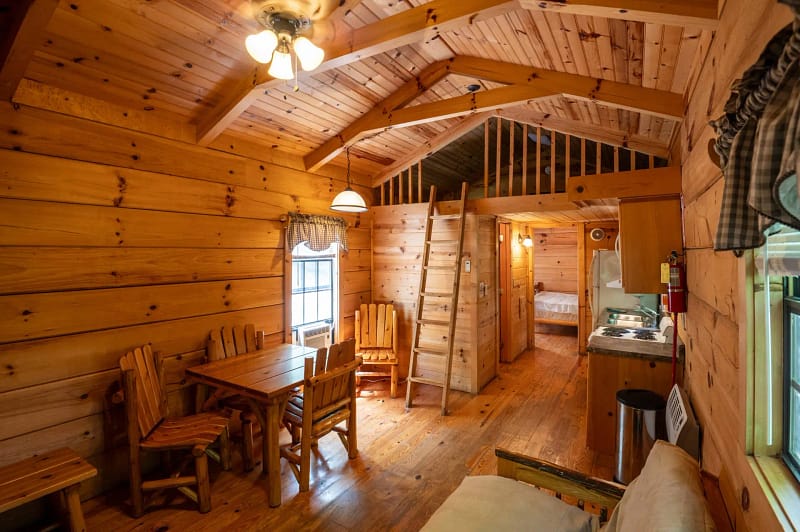 A cozy wooden cabin interior with a dining table and chairs on the left, a small kitchenette on the right, and a loft area accessed by a ladder. The ceiling features exposed beams, and warm lighting enhances the inviting atmosphere. A bed is visible in the back room.