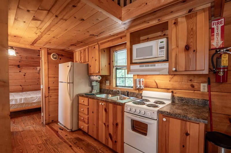 A cozy cabin kitchen with wooden walls and ceiling. The kitchen features a white refrigerator, stove, and microwave. There are shelves and cupboards, a sink, and kitchen tools. A fire extinguisher is mounted on the wall. A bedroom is visible in the background.