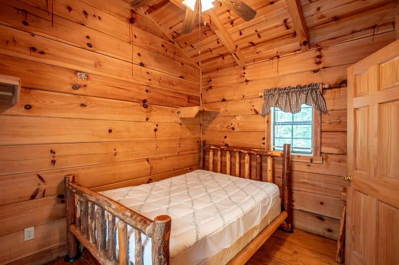 A cozy log cabin bedroom features wooden walls and a wooden ceiling with beams. The room is furnished with a rustic wooden bed frame and a mattress, under a simple window with a checkered valance curtain. A ceiling fan provides lighting overhead.