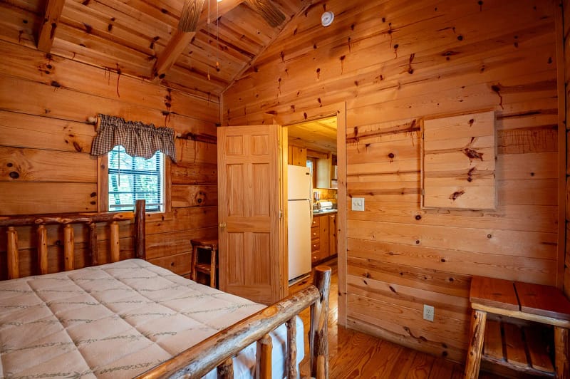 A cozy log cabin bedroom with wooden walls, ceiling, and furniture. A bed with a simple mattress is in the foreground. The door is open, revealing a view of the kitchen with a white refrigerator. A small window with checkered curtains is on the left.