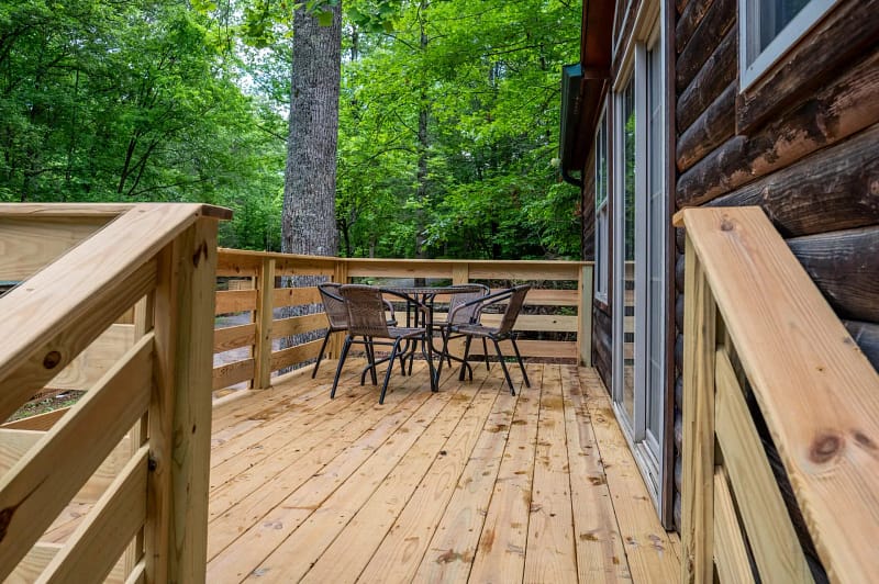 A wooden deck attached to a cabin in a forest, featuring a round black metal table and four wicker chairs. The deck is surrounded by a simple wooden railing, with lush green trees in the background. Sliders offer entry into the cabin.