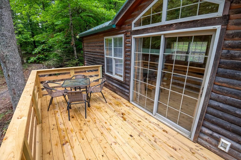 A wooden cabin with a spacious deck, featuring a glass-top table and four metal chairs. The deck overlooks a lush, green forest. Large sliding glass doors lead from the deck to the cabin's interior, and a window is visible next to the doors.