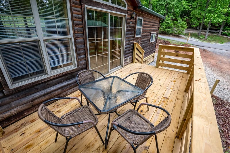 A wooden deck attached to a rustic cabin with large windows. The deck features a square glass table and four wicker chairs. Trees and a gravel pathway can be seen in the background. The setting appears peaceful and inviting.