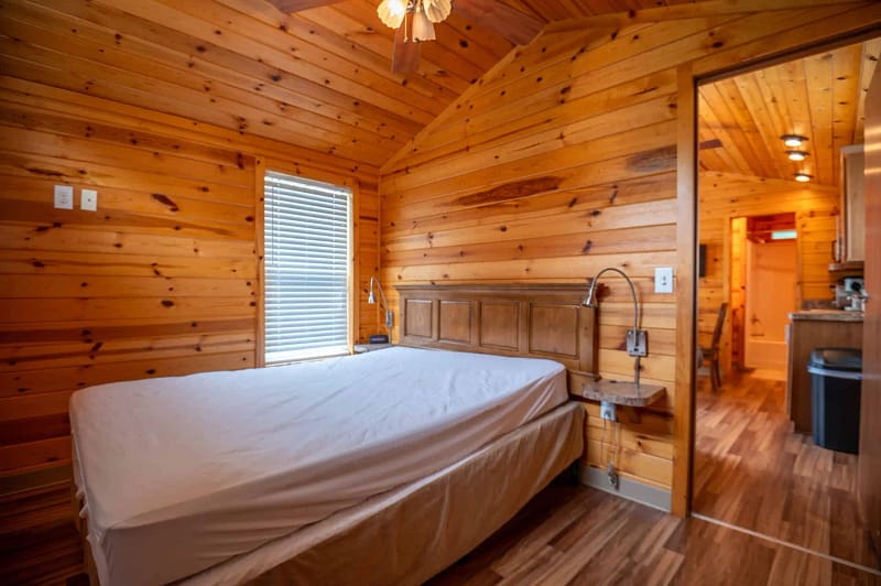 A cozy wooden cabin interior offers charming lodgings with a bedroom featuring wooden-paneled walls and ceiling. The room has a double bed with a simple headboard, a small window with blinds on the left, and a doorway leading to a kitchenette area in the background.