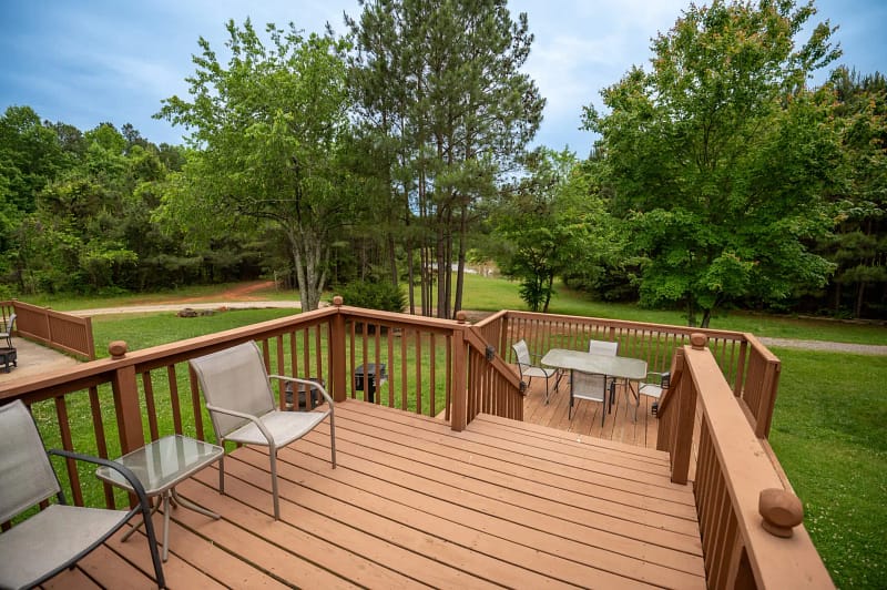 A wooden deck with outdoor furniture overlooks a lush green yard with trees. The deck has two chairs facing each other and a table with four chairs below on a lower section. This serene lodging extends into a dense forest, offering peaceful places to stay amidst nature.
