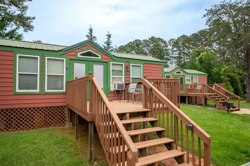 Three small, elevated cabins with reddish-brown exteriors and green trim, each with a wooden staircase and small porch. These cozy lodgings sit on a grassy area with tall trees in the background. The cabins, featuring multiple windows and white doors, offer charming places to stay under a partly cloudy sky.