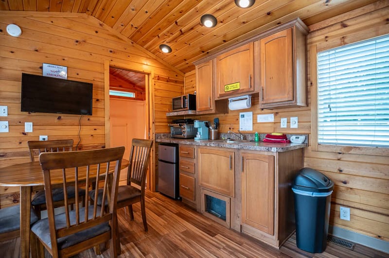 A small wooden kitchen and dining area with a round table and four chairs. The lodgings feature wooden cabinets, a sink, a microwave, a mini-fridge, and a coffeemaker. To the left is a flat-screen TV mounted on the wall, and a window with blinds is on the right.