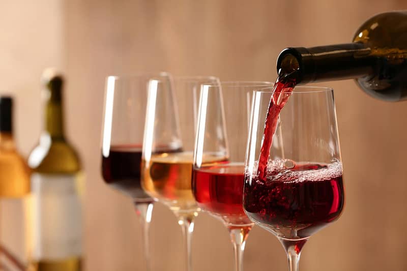 A bottle pours red wine into a glass, with three other wine glasses containing red, rose, and white wine lined up beside it.