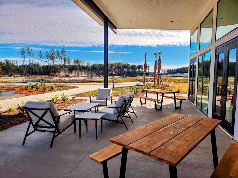 Outdoor patio with modern furniture under a sheltered roof, overlooking a scenic landscape with trees and open fields. Wooden picnic tables and cushioned seating arrangements are placed on the concrete floor, creating a serene and inviting atmosphere perfect for weekly gatherings or monthly events.